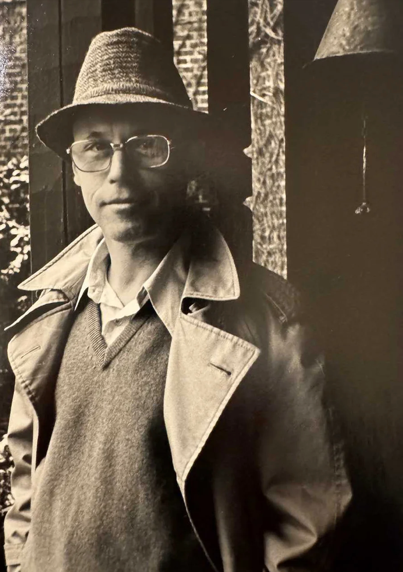 Artist Arthur Gebhardt as a young adult wearing a trench coat, hat, and glasses, looking stoically at the camera